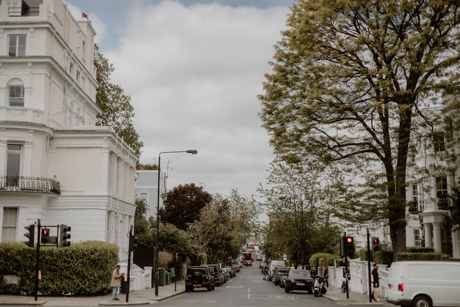 A street scene in Belgravia showing a residential area with white, ornate multi-story buildings on both sides, surrounded by green trees and bushes. In the foreground, women are waiting at pedestrian 