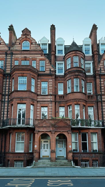 A multi-storey residential building constructed of red brick with decorative stone accents, featuring multiple large windows with white frames, some with small balconies enclosed by black metal railin