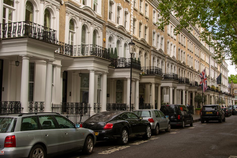 The image shows a row of elegant, white Victorian-style townhouses in Belgravia, each featuring ornate black wrought iron balconies, large bay windows, and decorative columns at the entrances. Parked along the street are several vehicles, including a silver estate car and a black sports car, situated on a paved road with designated parking spaces. A black van belonging to Man and Van Belgravia is visible, suggesting a home relocation or furniture transport process, with the van positioned near the building to facilitate loading or unloading. The scene is set during daylight hours, with leafy trees partially visible at the top right corner, and Union Jack flags hanging from some of the properties. The environment indicates a quiet, residential area typical of house removals and packing and moving activities, with the vehicles and equipment aligned for efficient house removals in this upscale Belgravia neighbourhood.