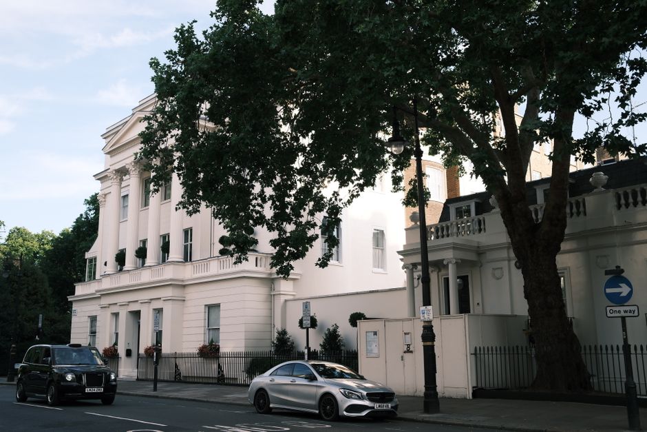 The image shows a row of elegant, white Victorian-style townhouses in Belgravia, each featuring ornate black wrought iron balconies, large bay windows, and decorative columns at the entrances. Parked along the street are several vehicles, including a silver estate car and a black sports car, situated on a paved road with designated parking spaces. A black van belonging to Man and Van Belgravia is visible, suggesting a home relocation or furniture transport process, with the van positioned near the building to facilitate loading or unloading. The scene is set during daylight hours, with leafy trees partially visible at the top right corner, and Union Jack flags hanging from some of the properties. The environment indicates a quiet, residential area typical of house removals and packing and moving activities, with the vehicles and equipment aligned for efficient house removals in this upscale Belgravia neighbourhood.